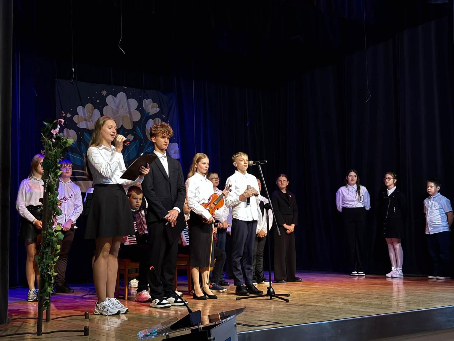 Uczniowie w białych koszulach na scenie Gminnego Ośrodka Kultury w Podegrodziu podczas programu artystycznego z okazji Dnia Edukacji Narodowej, w tle muzycy ze skrzypcami i akordeonem