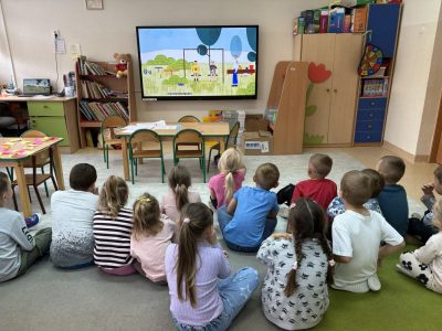 Dzieci siedzą na dywanie w sali SP w Olszanie i oglądają film edukacyjny na ekranie podczas zajęć
