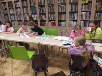 Dzieci przy stołach z otwartymi książkami w Bibliotece Gminnej w Podegrodziu podczas Nocy Bibliotek, w tle regały z książkami