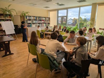 Uczniowie przy stołach w czytelni Biblioteki Gminnej w Podegrodziu słuchają bibliotekarki stojącej przy tablicy, w tle regały z książkami