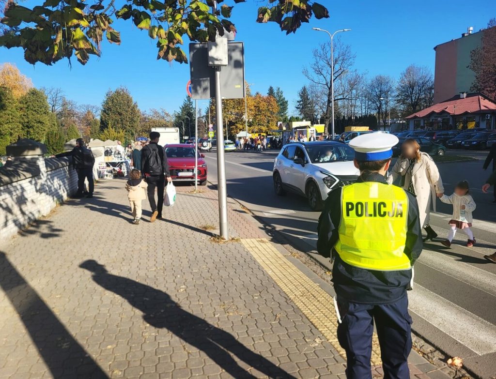 Policjantka ruchu drogowego w odblaskowej kamizelce przy przejściu dla pieszych obok cmentarza, po chodniku idą odwiedzający